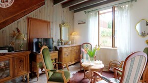A cozy living room with a wooden staircase, a television, a green armchair, a wooden cabinet, and a window with curtains. - Charming Cottage with Terrace and Garden, Pets Welcome (Bussières)