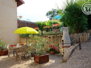 Eine Terrasse im Freien mit Tisch und Stühlen, einem gelben Sonnenschirm, Kübelpflanzen und einer Steinsitzbank.