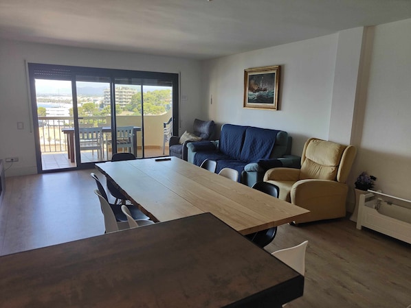 Un soggiorno moderno con un grande tavolo in legno, un divano e una poltrona. È presente un balcone con vista sulla città.
