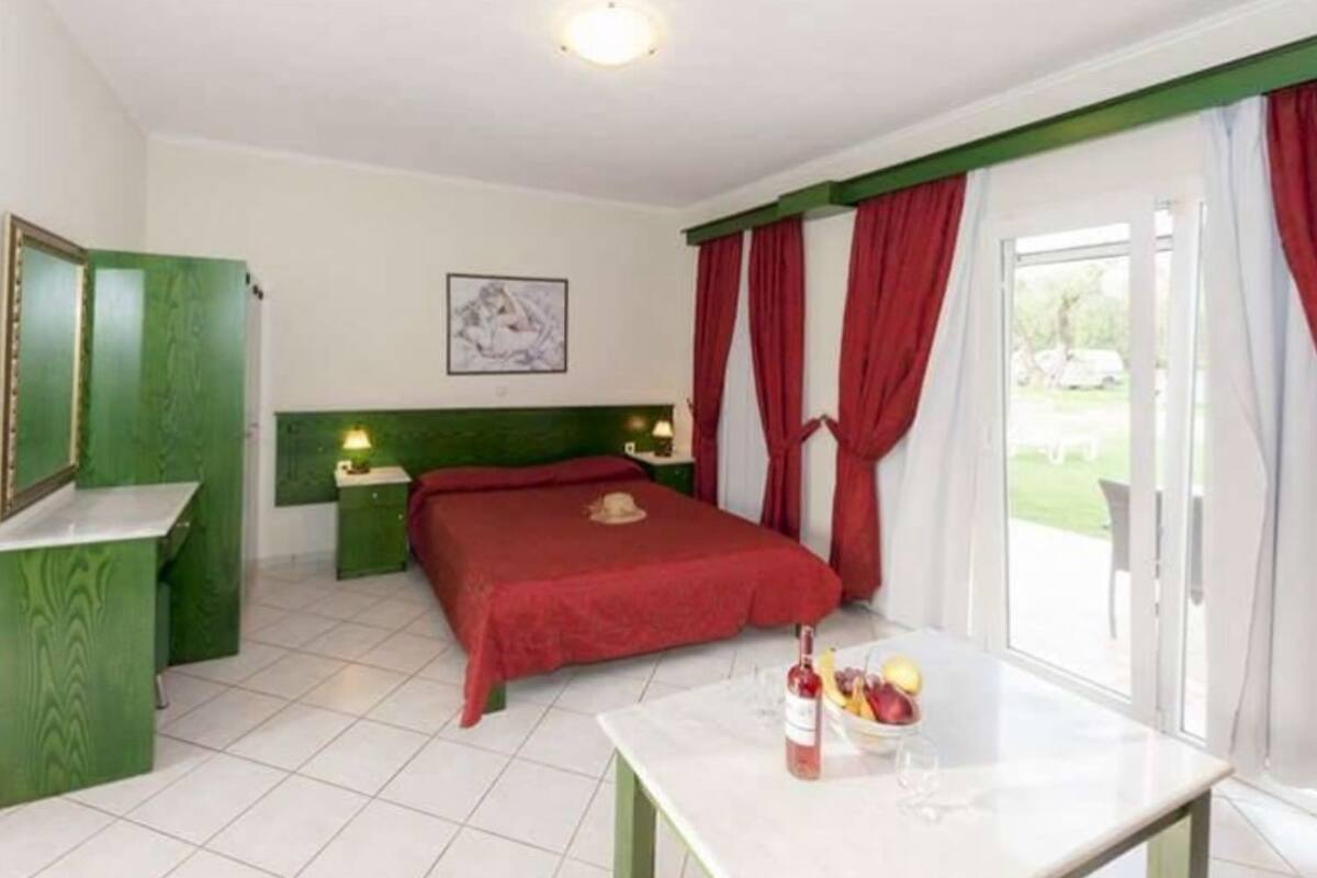 A hotel room with a bed, a table with a fruit bowl, green cabinets, and a window with red curtains.