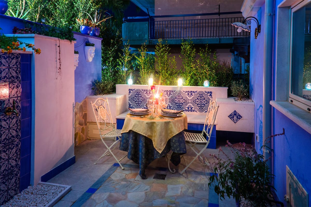 Un espace de restauration en plein air avec une table dressée pour deux personnes, entouré de murs aux motifs bleus et blancs, et un balcon situé au-dessus.