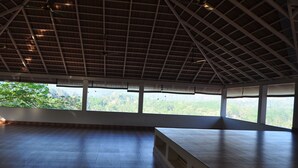 A spacious room with a high ceiling, large windows, and a view of greenery outside. - INDIMASI - Ayurveda & Healing Village (Nedumangad)
