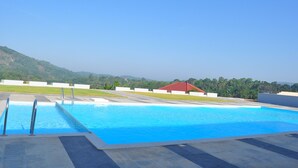 A swimming pool with a clear view of the surrounding landscape, including a building with a red roof and a hilly backdrop. - INDIMASI - Ayurveda & Healing Village (Nedumangad)