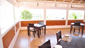A dining area with brick walls, large windows, and wooden tables and chairs. - INDIMASI - Ayurveda & Healing Village (Nedumangad)