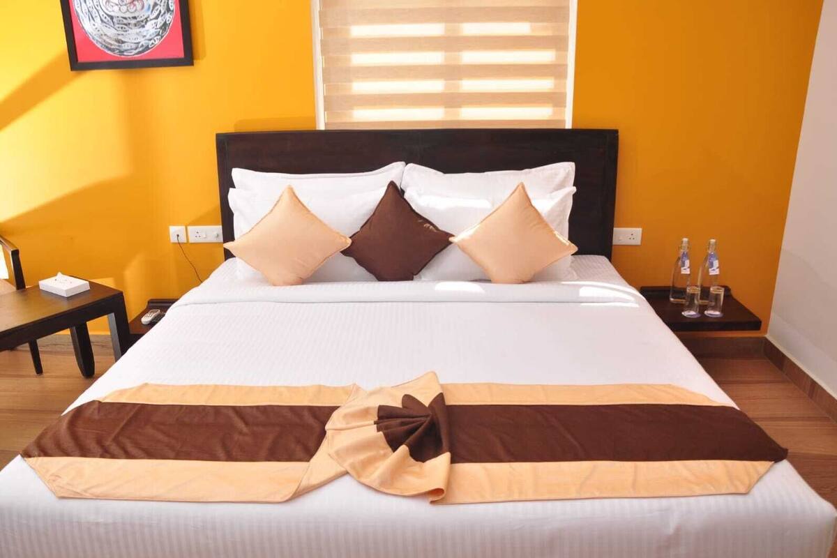 A neatly made bed with a brown and beige blanket, a wooden nightstand with a lamp and a bottle, and a chair in a room with yellow walls and a window with blinds.