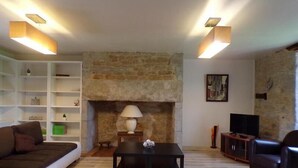 A living room with a stone fireplace, a sofa, a coffee table, and a TV. - Gîte Le Chant des Ifs (Bio)