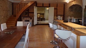 A kitchen with a wooden staircase, a dining area with white chairs, and a bar with stools. - Gîte Le Chant des Ifs (Bio)