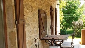 An outdoor patio with a stone wall, wooden shutters, and a tiled floor. - Gîte Le Chant des Ifs (Bio)
