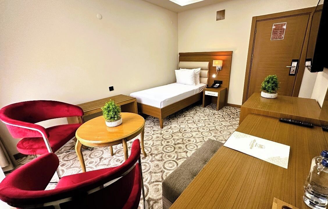 A hotel room with a bed, a desk, a chair, a small table with a plant, and a water bottle.