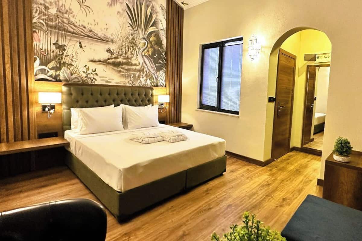 A hotel room with a large bed, a wooden floor, a mural on the wall, and a small potted plant.