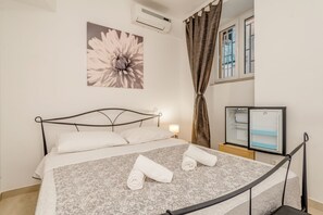 A neatly made bed with a patterned bedspread and two rolled towels, a wall-mounted air conditioner, a window with curtains, and a mini-fridge. - Porta Adriatica (Split)
