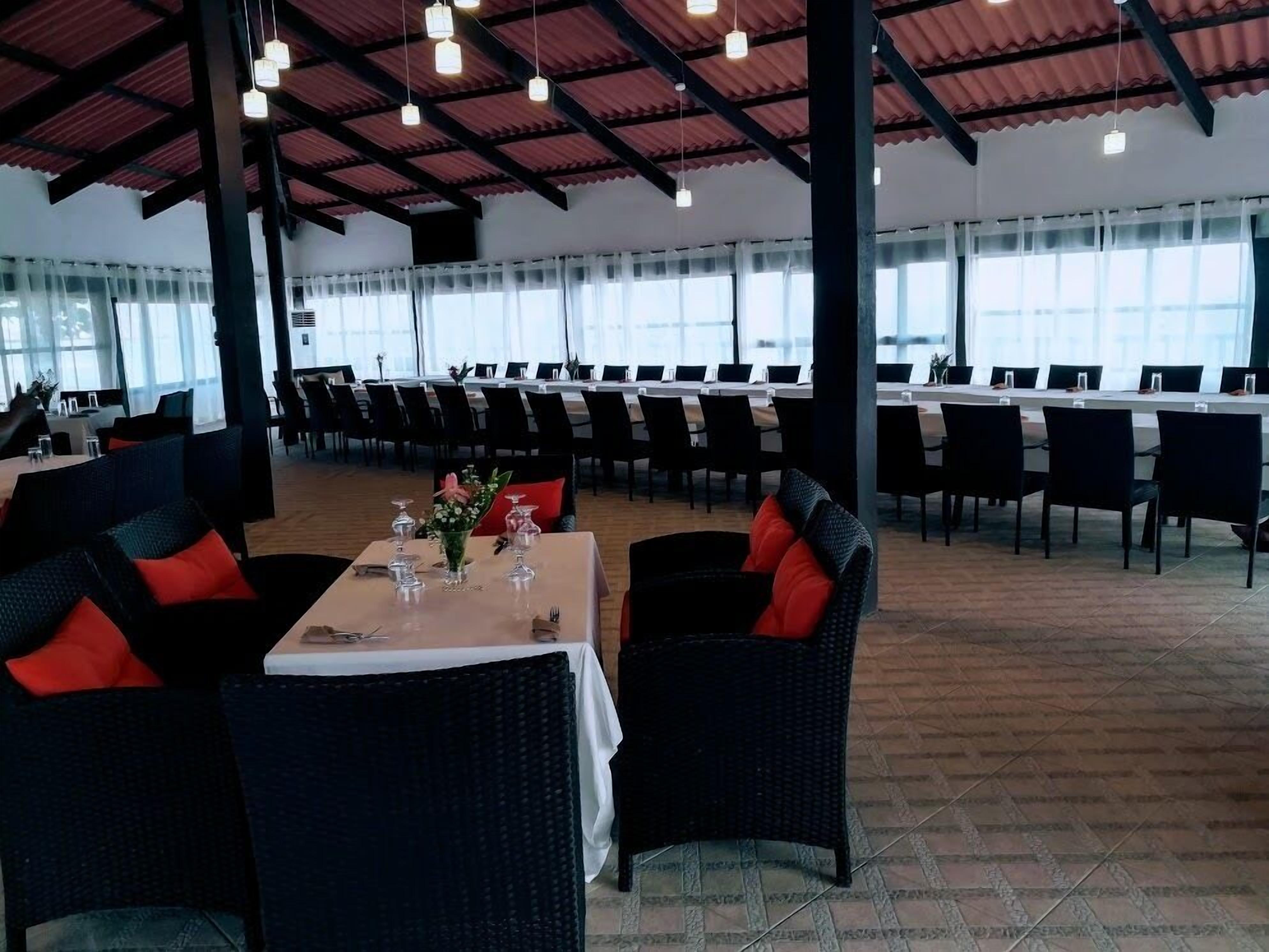 Un'ampia sala da pranzo con soffitti alti e ampie finestre, con tavoli apparecchiati e sedie in vimini con cuscini rossi.