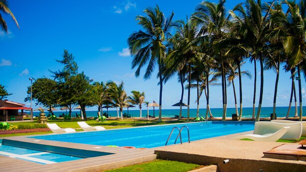 A pool area with lounge chairs, palm trees, and a view of the ocean. - Hotel Riviera Ramatou Plage (Lomé)