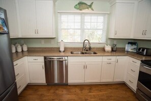 A kitchen with white cabinets, a stainless steel dishwasher, a window with blinds, and a fish decoration. - New Owner offering 20%rent discount for 7/28-8/4, 2028 Ramp Access-No Stairs (Duck)