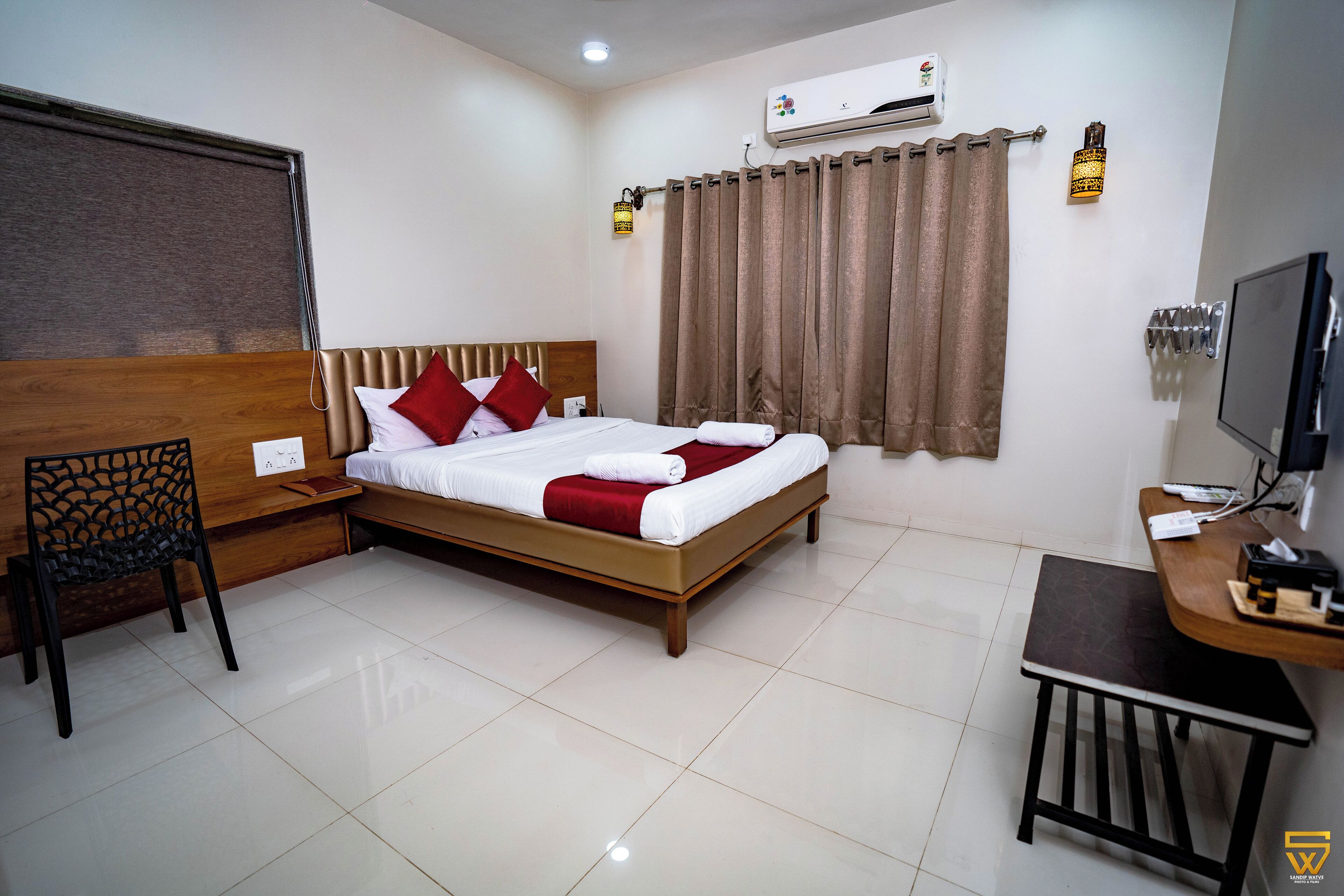 A hotel room with a bed, a desk, a chair, an air conditioning unit, and a television.