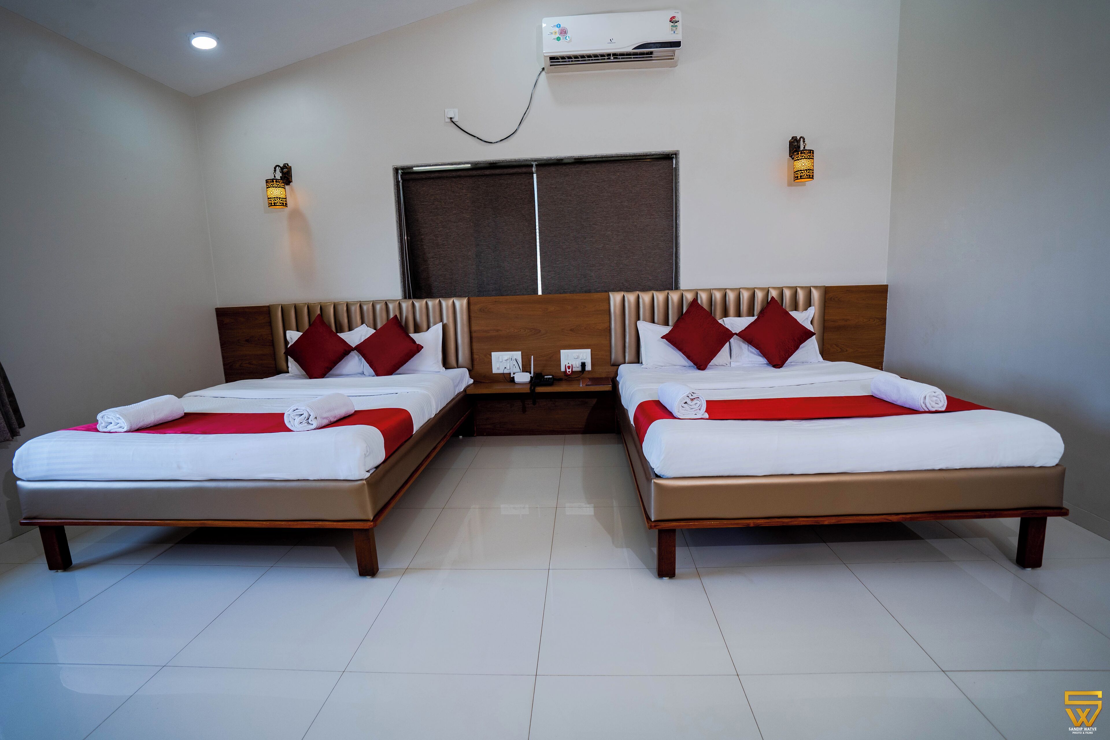 A hotel room with two beds, each with a wooden headboard, white bedding, and red accents. There is a window with a view, an air conditioning unit, and wall-mounted lights.