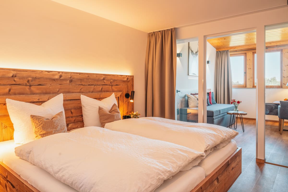 Ein modernes Hotelzimmer mit einem Holzkopfteil, einem großen Bett mit weißer Bettwäsche, einem kleinen Tisch mit Blumenvase und Blick auf die Stadt durch große Fenster.