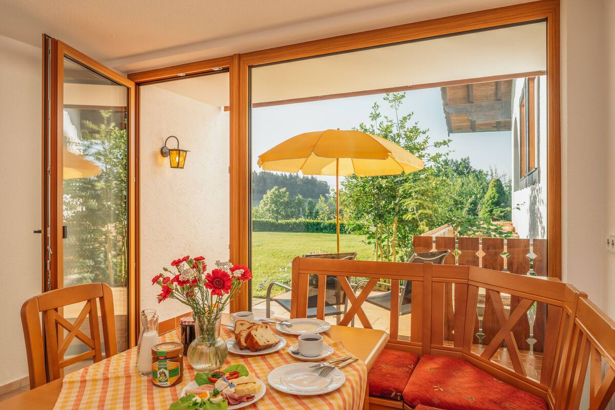 Ein Essbereich mit Tisch für zwei Personen, einer Blumenvase und Blick auf einen Garten durch eine Glastür.
