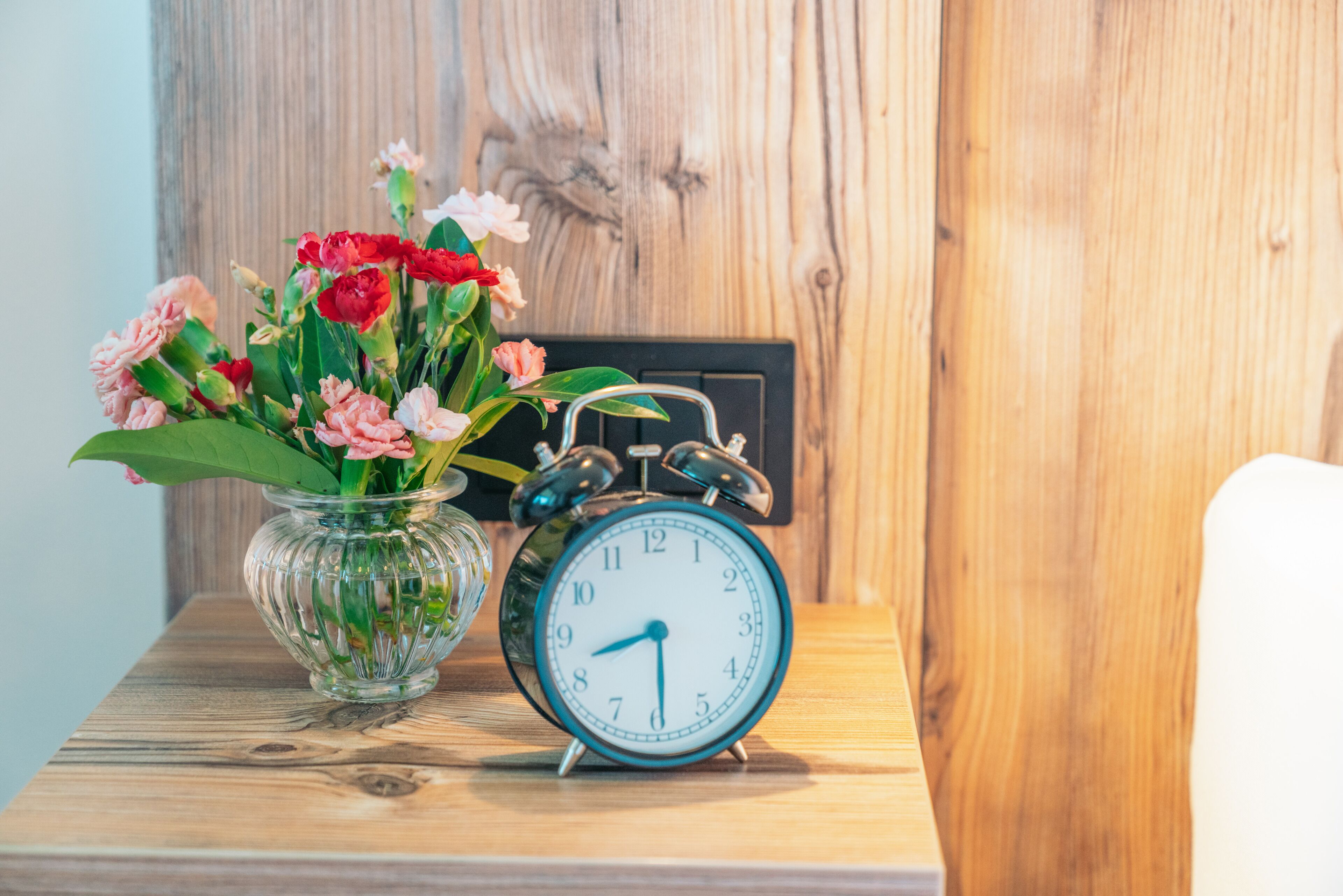 Une table en bois sur laquelle est posé un vase en verre transparent contenant des fleurs roses et rouges, ainsi qu’un réveil noir classique.