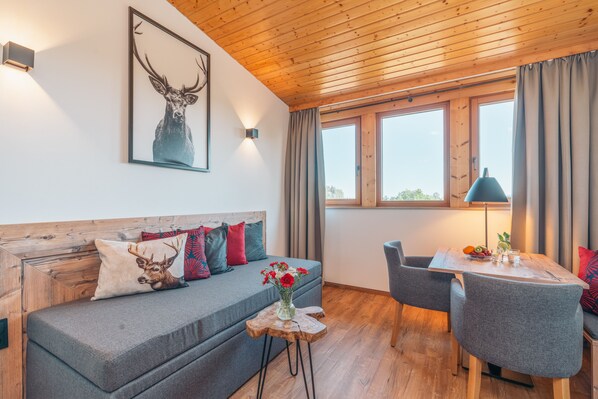 Una habitación acogedora con techo de madera, un sofá gris con cojines decorativos, una pequeña mesa de madera con un florero de flores, y un comedor con sillas y una mesa.