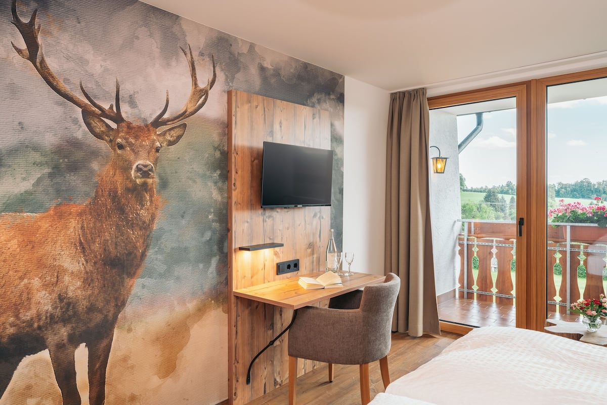 Ein Hotelzimmer mit einem großen Hirschgemälde, einem Holztisch mit Flachbildfernseher, einem Sessel, einem Bett und einem Balkon mit Blick auf Grünflächen.