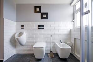 A modern bathroom with a white toilet, bidet, and a urinal. There are two wall-mounted light switches and a window to the right. - Lakovich Sea View Apartments (Rovinj)