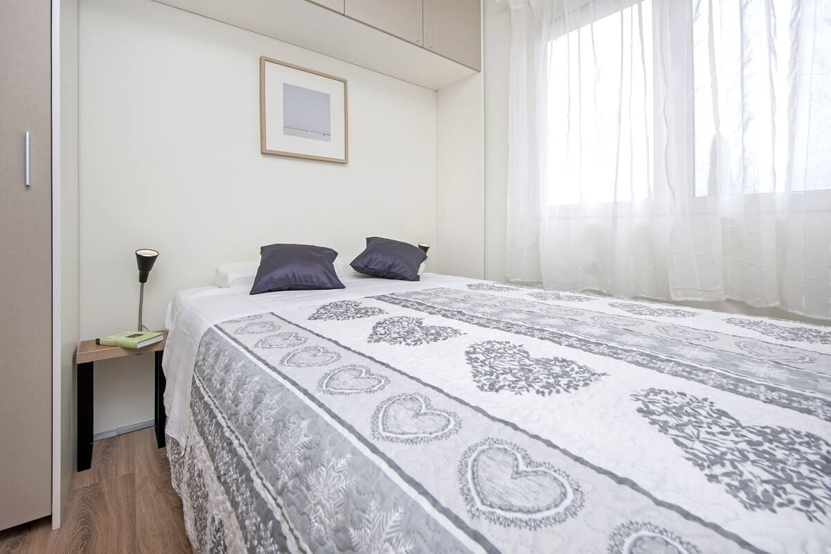 Un dormitorio con cama, mesita de noche, lámpara, un cuadro enmarcado en la pared y cortinas traslúcidas en la ventana.
