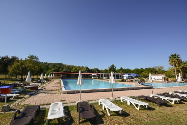 A pool area with lounge chairs and umbrellas, surrounded by trees and a building in the background. - Garden River - Campsite (Altidona)