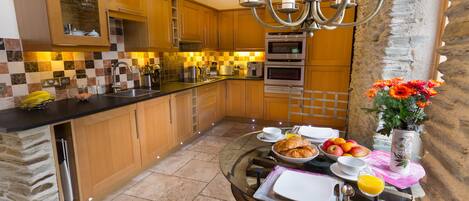 A modern kitchen with wooden cabinets, a stone accent wall, and a breakfast setup on a glass table.