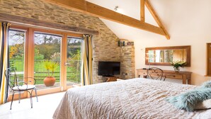 A bedroom with a large bed, a television, a dresser, and a balcony with a chair and a potted plant. - Coriander Cottages (Fowey)