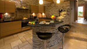 A modern kitchen with a stone island, bar stools, and a view of the outdoors. - Coriander Cottages (Fowey)