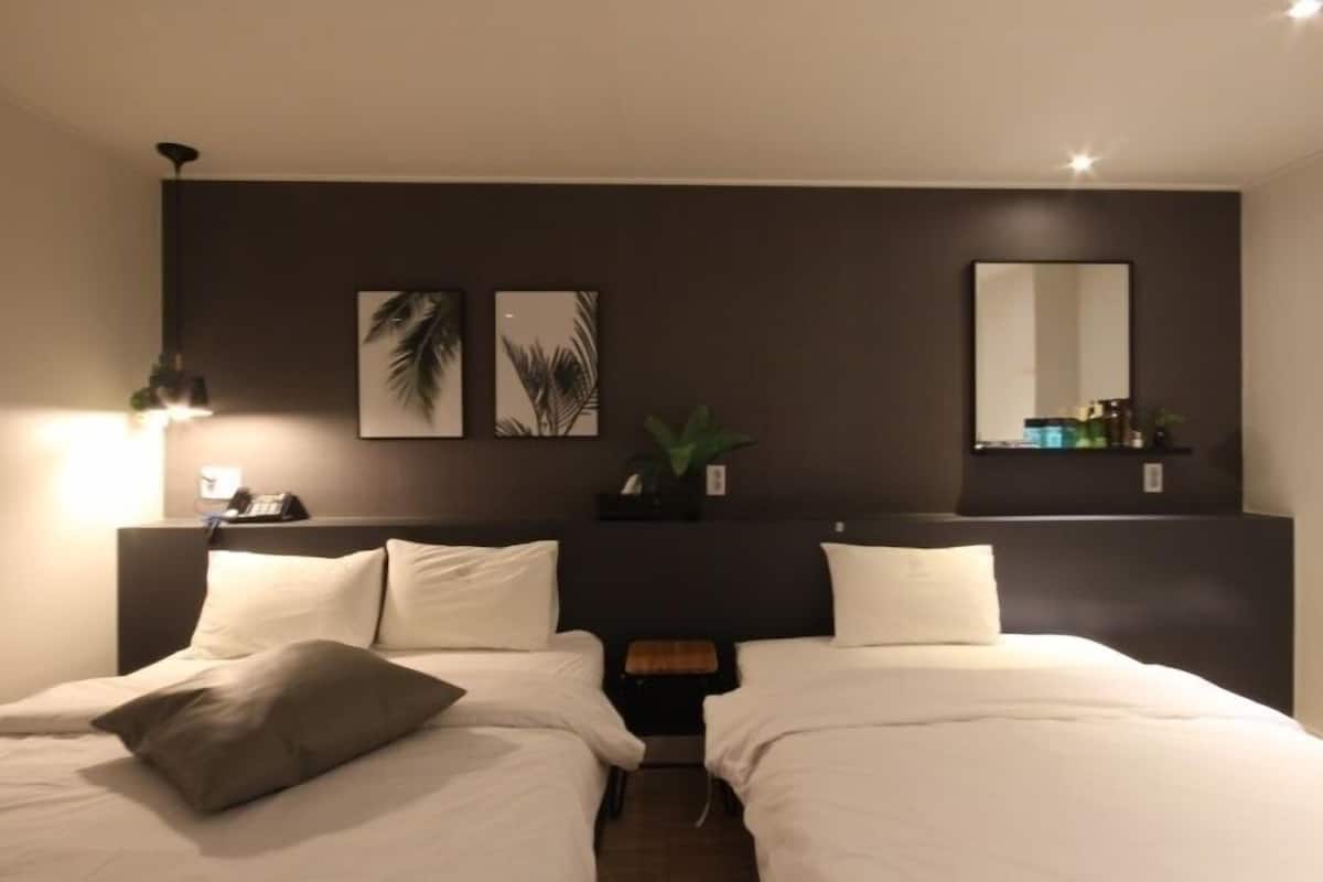 Una habitación de hotel con dos camas, una mesita de noche con lámpara, una pequeña mesa y una obra de arte en la pared.