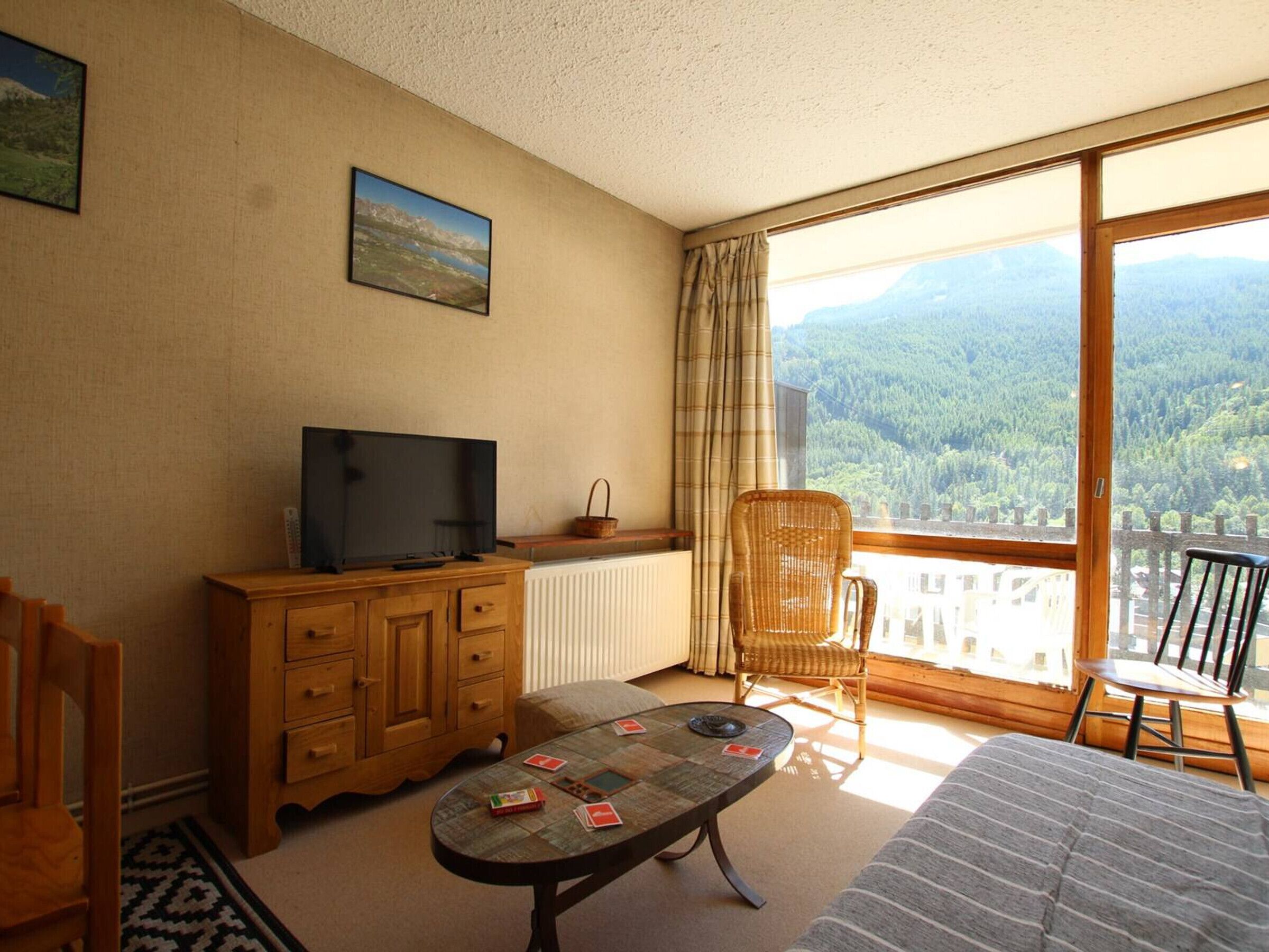 Een kamer met een houten kast, een televisie, een tafel met speelkaarten, een rieten stoel en een uitzicht op een bergachtig landschap door een groot raam.