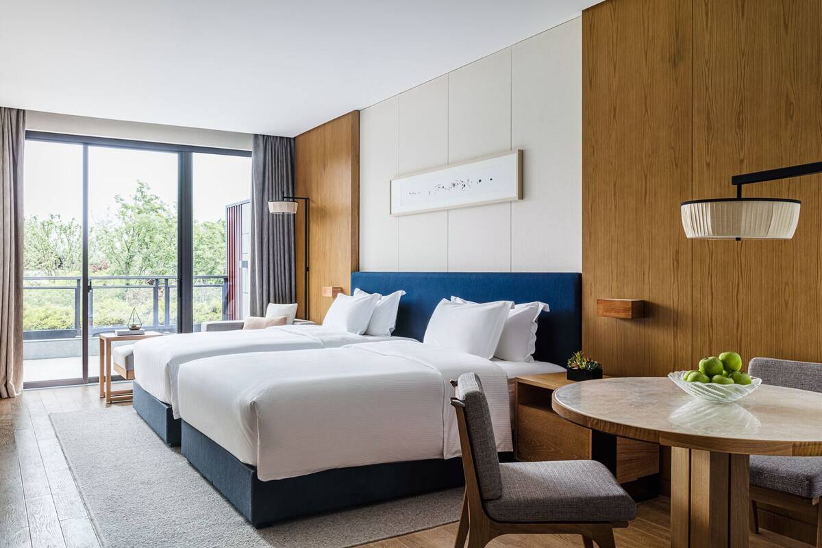 A modern hotel room with a large bed, a dining table, and a balcony with a view of greenery.