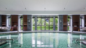 A large indoor swimming pool with a view of greenery outside, surrounded by wooden paneling and wicker furniture.