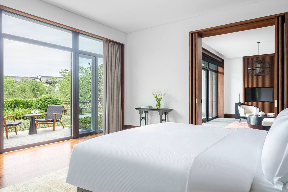 A modern hotel room with a large bed, a small table with a vase, a TV, and a view of the garden and building outside.