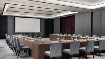 Una sala conferenze con lunghe tavole, sedie grigie e uno schermo per proiettore.