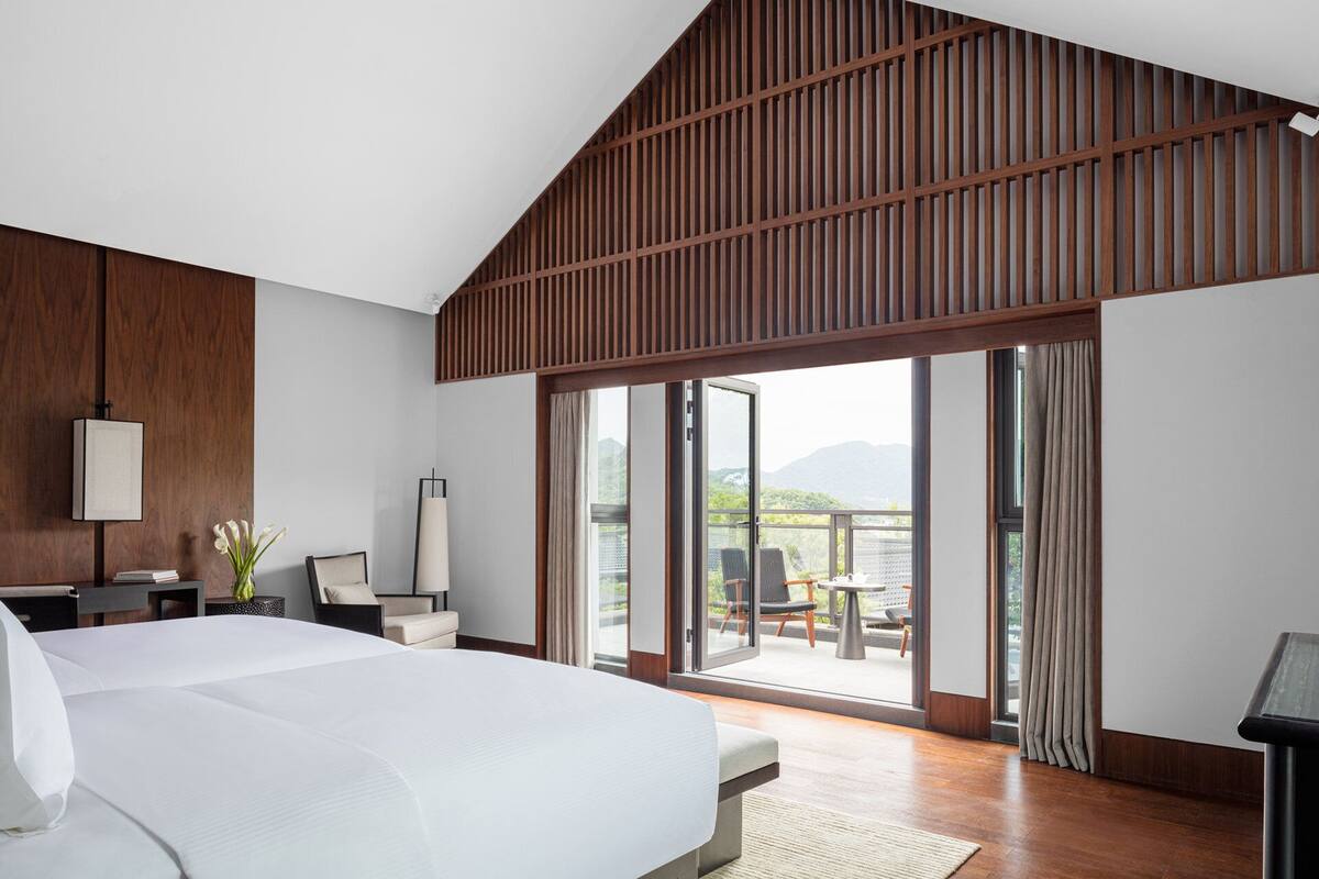 A modern hotel room with a large bed, a desk, and a view of a balcony with outdoor seating.