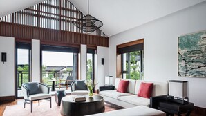 A modern living room with a sofa, chairs, a coffee table, and a TV stand. Large windows provide natural light and views of the outdoors.