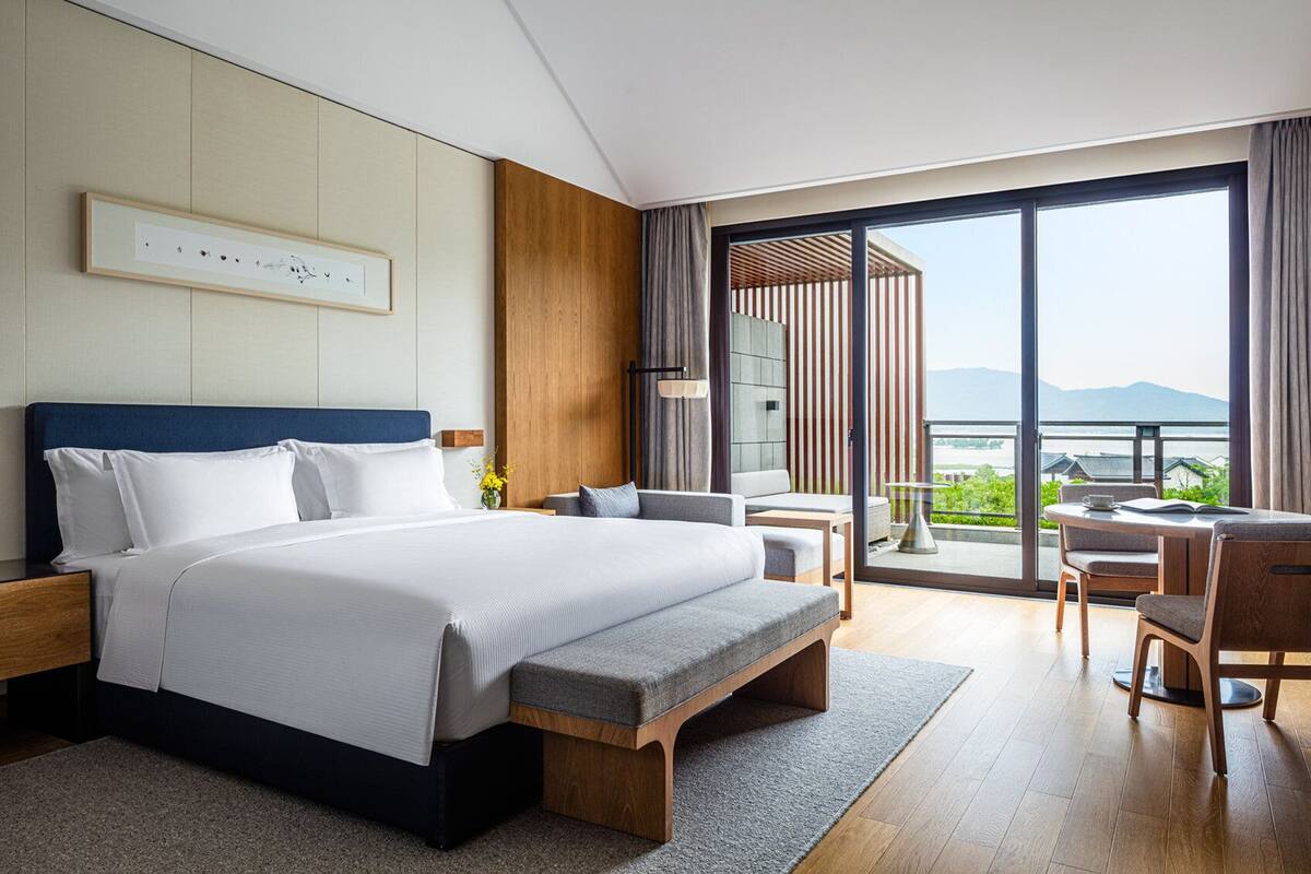 A modern hotel room with a large bed, a seating area with a table, and a balcony with a view of the mountains.
