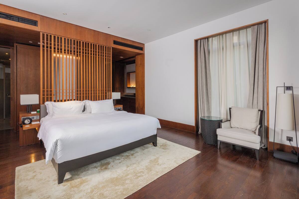 A modern hotel room with a large bed, a chair, a bedside table, and a lamp.