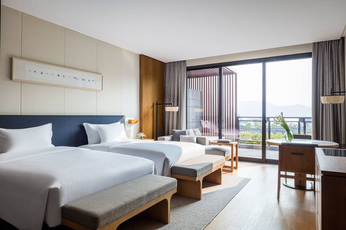 A hotel room with two beds, a desk, and a balcony with a view.