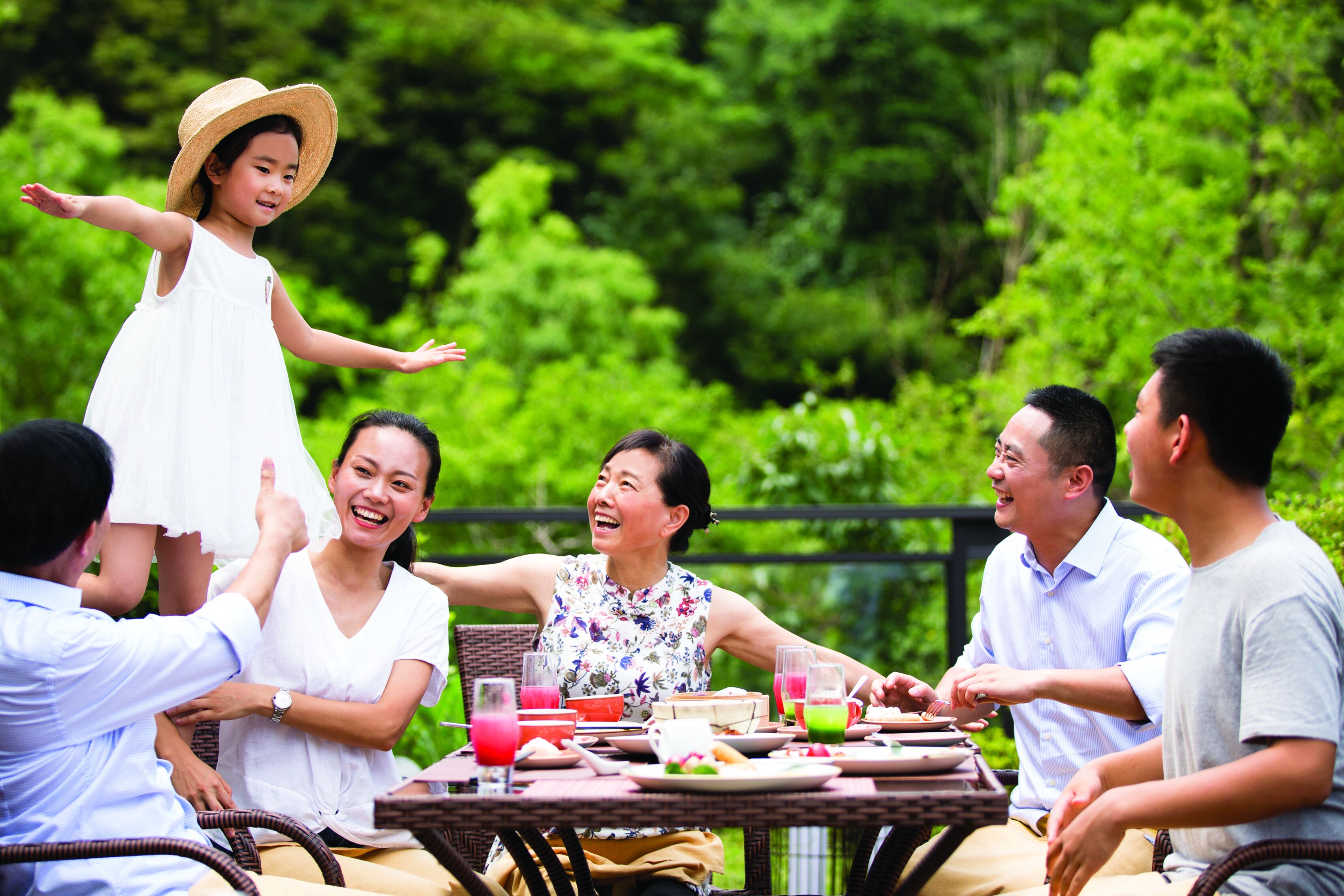 A group of people seated around a table outdoors with greenery in the background.