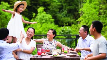 A group of people seated around a table outdoors with greenery in the background.