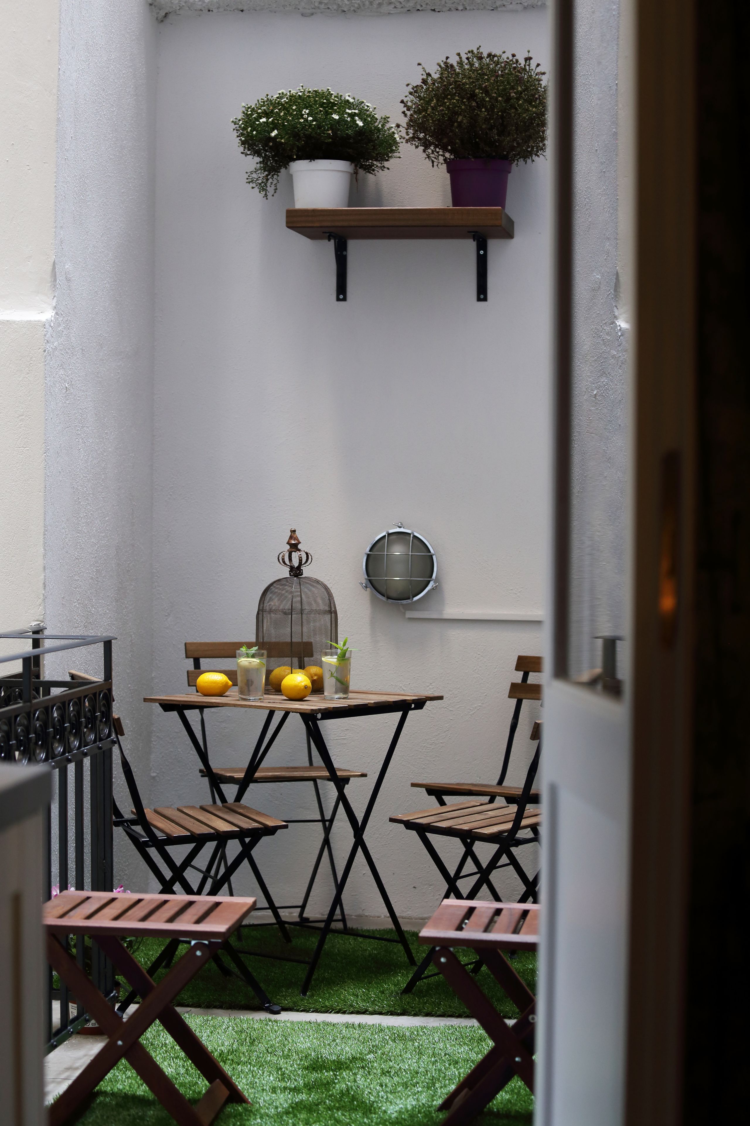 En liten sittplats utomhus med ett bord och stolar, en fågelbur och krukväxter mot en vit vägg.