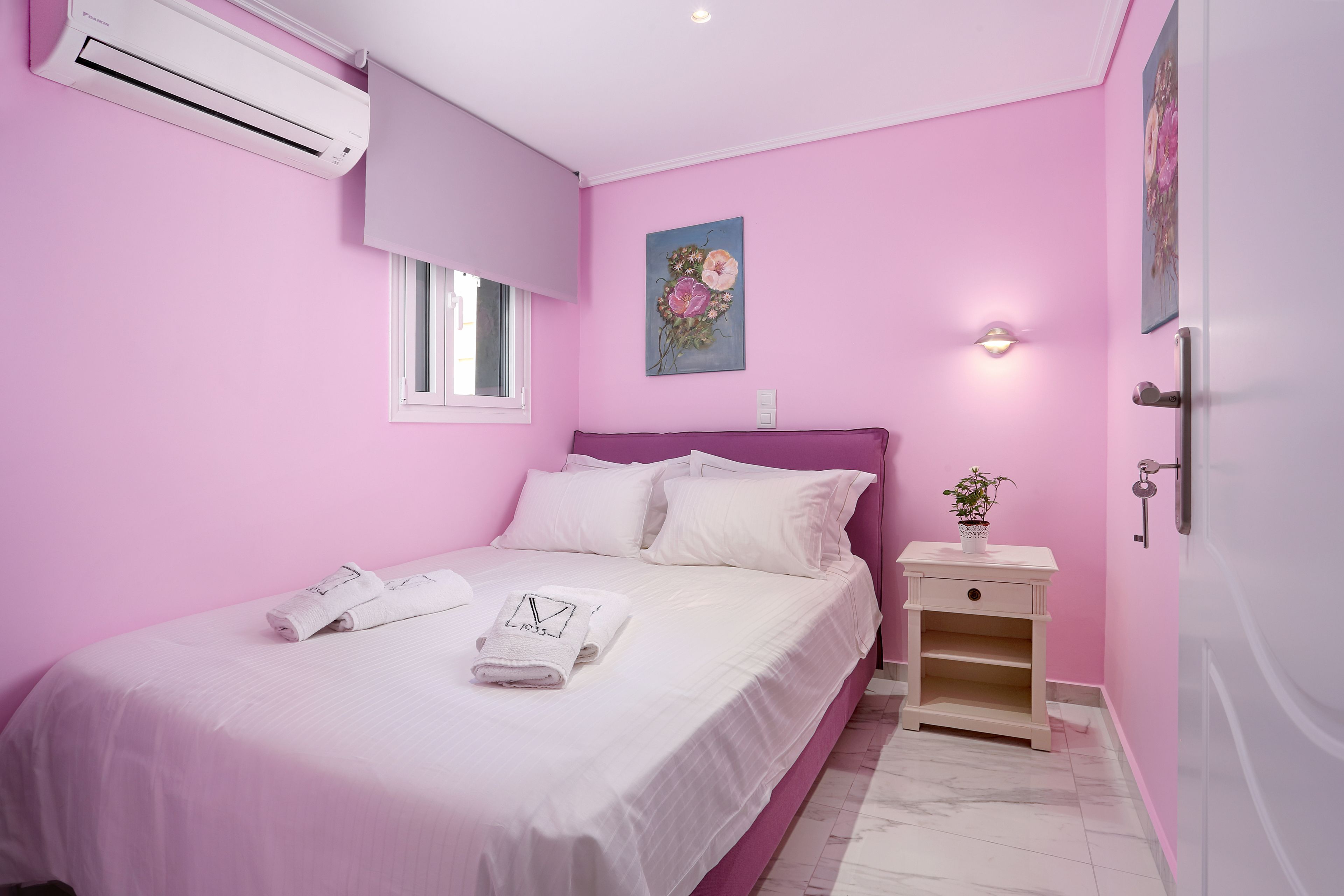 A pink-themed bedroom with a bed, white towels, a nightstand, and a wall-mounted air conditioner.