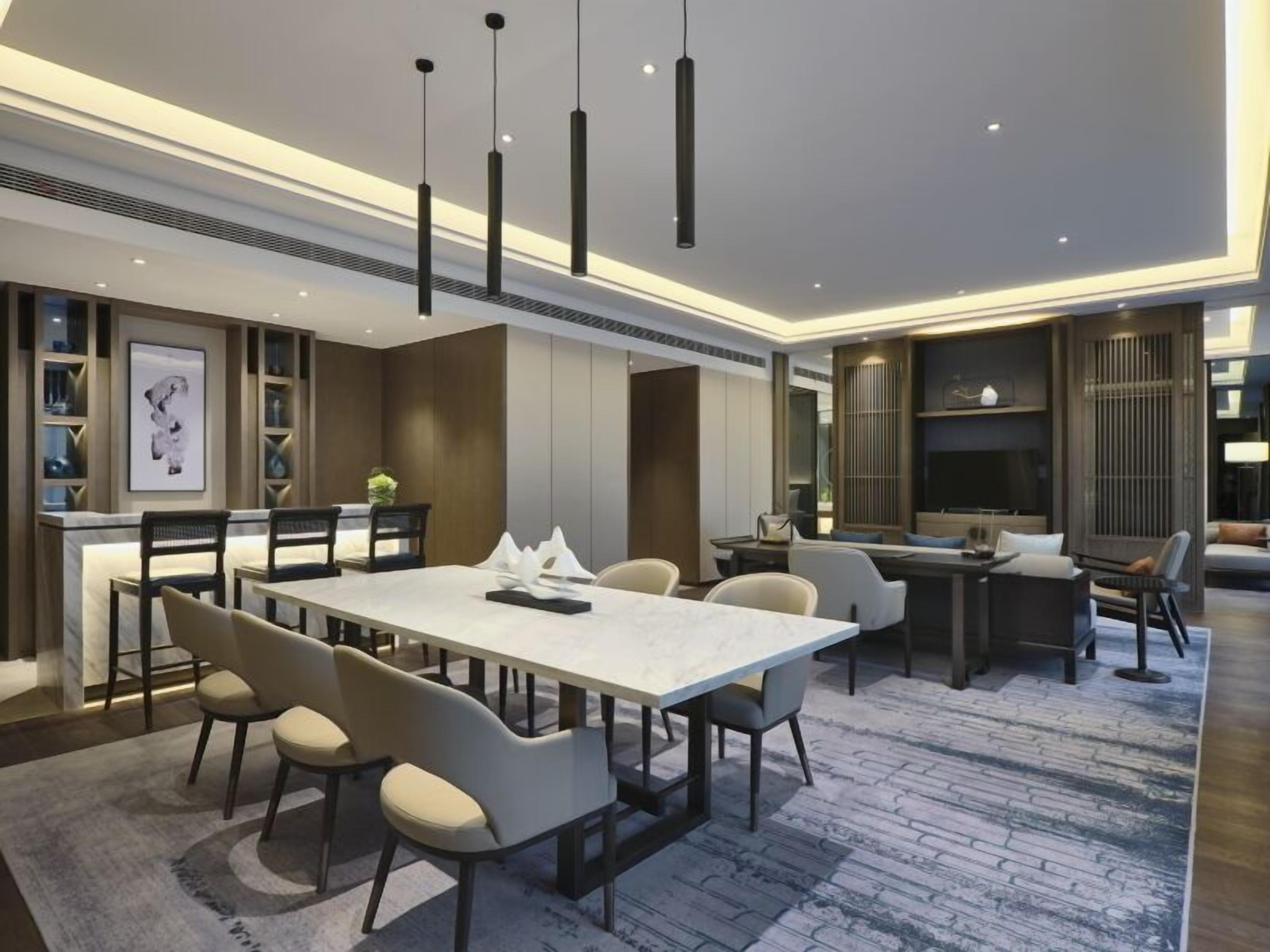 A modern hotel lobby with a central dining area, seating arrangements, and a TV screen.