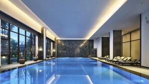Une piscine intérieure moderne avec une eau d’un bleu cristallin, des chaises longues et de grandes fenêtres offrant une vue sur l’extérieur.
