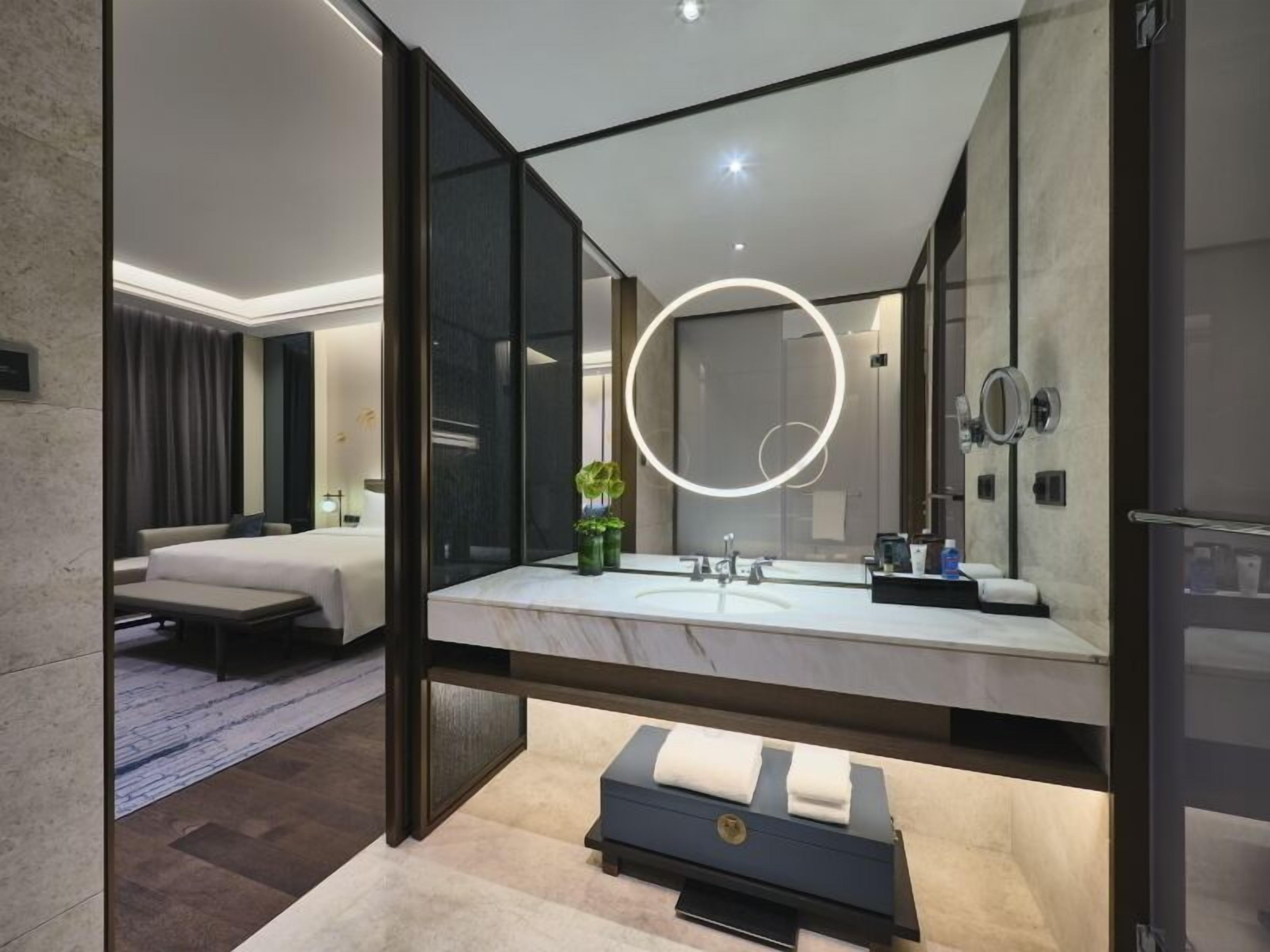 Een moderne hotelbadkamer met een grote spiegel, een ronde lamp en een bankje met handdoeken.