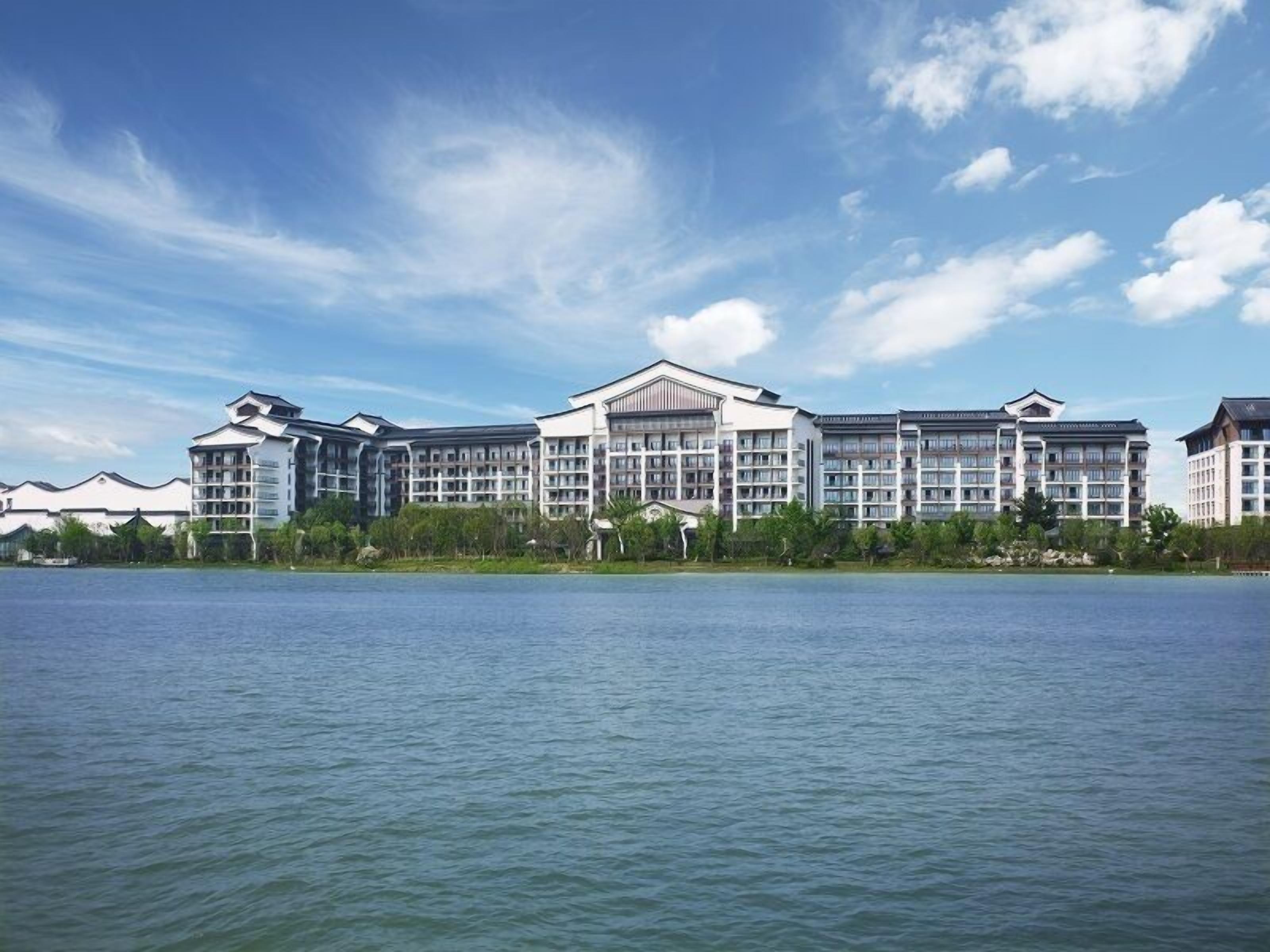 A hotel with multiple buildings, a large body of water in the foreground, and a clear sky.